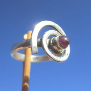 Könnte beinhalten: Silberring mit Spiraldesign und einem kleinen, runden, purpurroten Edelstein. Der Ring ist vor einem klaren blauen Himmel platziert, was seine einzigartige Form und Farbe hervorhebt.