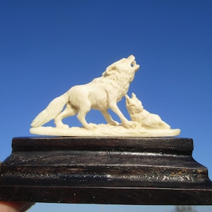 Könnte beinhalten: Eine geschnitzte, elfenbeinfarbene Skulptur von zwei Wölfen. Ein Wolf steht und heult, während der andere liegt. Die Skulptur ist auf einem dunkelbraunen Holzsockel montiert. Der Hintergrund ist ein klarer blauer Himmel.