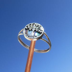 May include: A silver ring with a tree of life design. The ring has a round face with an openwork tree motif and iridescent inlay. The band splits into two delicate strands. The ring is held against a bright blue sky.