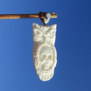 Könnte beinhalten: Ein geschnitzter cremefarbener Anhänger, der eine Eule mit einem in die Brust integrierten Totenkopf darstellt. Der Anhänger ist an einer silberfarbenen Metallöse mit einem kleinen, runden, schillernden Steineinsatz befestigt.