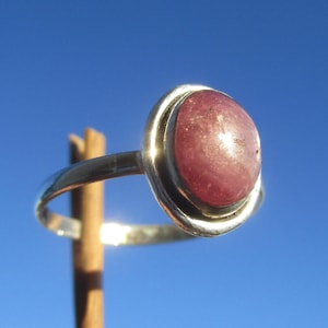 Pode incluir: Um anel de prata com uma pedra preciosa lisa de formato oval na cor rosa empoeirado, engastada em um bisel de metal polido. A banda é fina e circular, reflete a luz em sua superfície metálica e apresenta um design simples com pedra rosa.