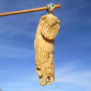 以下が含まれることがあります： 薄いベージュ色の彫刻ペンダント。木または骨で作られている可能性があり、様式化された動物の姿を描いています。 ペンダントは、小さな緑色の宝石が付いた銀色のベイルから吊り下げられています。 背景は澄んだ青い空です。