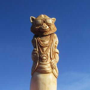 Könnte beinhalten: Eine hellfarbene Holzskulptur eines Affen mit Sonnenbrille, Weste und geschichteten Halsketten. Der Affe hat ein strukturiertes Fell und ist mit detaillierten Gesichtszügen, einschließlich Ohren und Nase, vor einem klaren blauen Himmel geschnitzt.