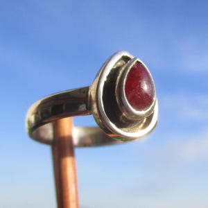 Op de afbeelding: Een zilveren ring met een traanvormige, dieprode edelsteen gezet in een gladde, gepolijste metalen zetting. De ringband is van massief zilver met een reflecterende afwerking en heeft een strak, eenvoudig ontwerp dat de steen accentueert.