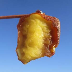 Puede incluir: Un trozo de ámbar con una cara tallada, sostenido por un palillo de madera. El ámbar es una mezcla de amarillo y marrón, con una pequeña gema roja engastada en plata en la parte superior. El fondo es un cielo azul claro.
