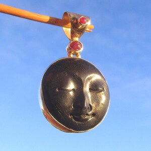 Puede incluir: Un colgante circular que presenta un rostro tallado en piedra negra con los ojos cerrados y una sonrisa sutil. El colgante está engastado en un marco de metal dorado e incluye una reasa adornada con dos pequeñas piedras rojas redondas.