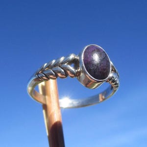 Könnte beinhalten: Ein Silberring mit einem polierten, ovalen violetten Stein in einer Fassung. Die Ringschiene weist ein strukturiertes, geflochtenes Muster auf, das an Weizenähren erinnert. Das Metall hat eine glatte, reflektierende Oberfläche im blauen Licht.