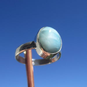 Op de afbeelding: Een zilveren ring met een grote, ovale, lichtblauwe edelsteen. De steen heeft een gemarmerd uiterlijk met witte patronen. De ring is geplaatst tegen een heldere blauwe lucht, wat de kleur en het ontwerp van de edelsteen benadrukt.
