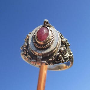 May include: A silver locket ring featuring an oval-shaped, cabochon-cut reddish-pink stone set in a twisted metal border. The ring band and setting are decorated with intricate metal scrollwork, small spheres, and a visible hinge mechanism.
