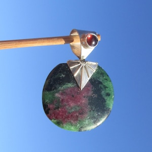 Könnte beinhalten: Ein runder Anhänger mit einem gesprenkelten grün-dunkelrosa Stein, gefasst in eine silberfarbene Metallfassung mit Sonnenstrahlen-Design. Die Öse ist mit einem kleinen, runden, dunkelroten Edelstein verziert und hängt an einem Holzstab.