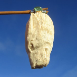 May include: A carved, off-white pendant in the shape of an animal head, possibly a wolf or dog, hangs from a wooden stick. A small, green gemstone is set in silver at the top. The background is a clear, bright blue sky.