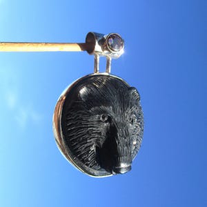 Könnte beinhalten: Silberanhänger mit einem geschnitzten schwarzen Bärenkopf-Design. Der Anhänger hat eine runde Form und ist an einer silbernen Öse mit einem kleinen, runden, irisierenden Edelstein aufgehängt. Der Hintergrund ist ein klarer blauer Himmel.