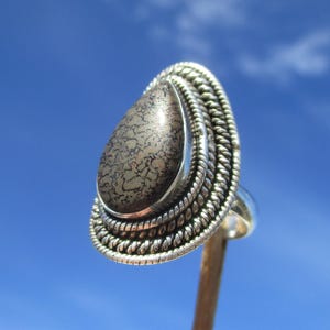 Pode incluir: Um anel de prata com uma pedra em forma de lágrima, de cor castanho e dourado, salpicada, engastada num desenho de filigrana de prata.