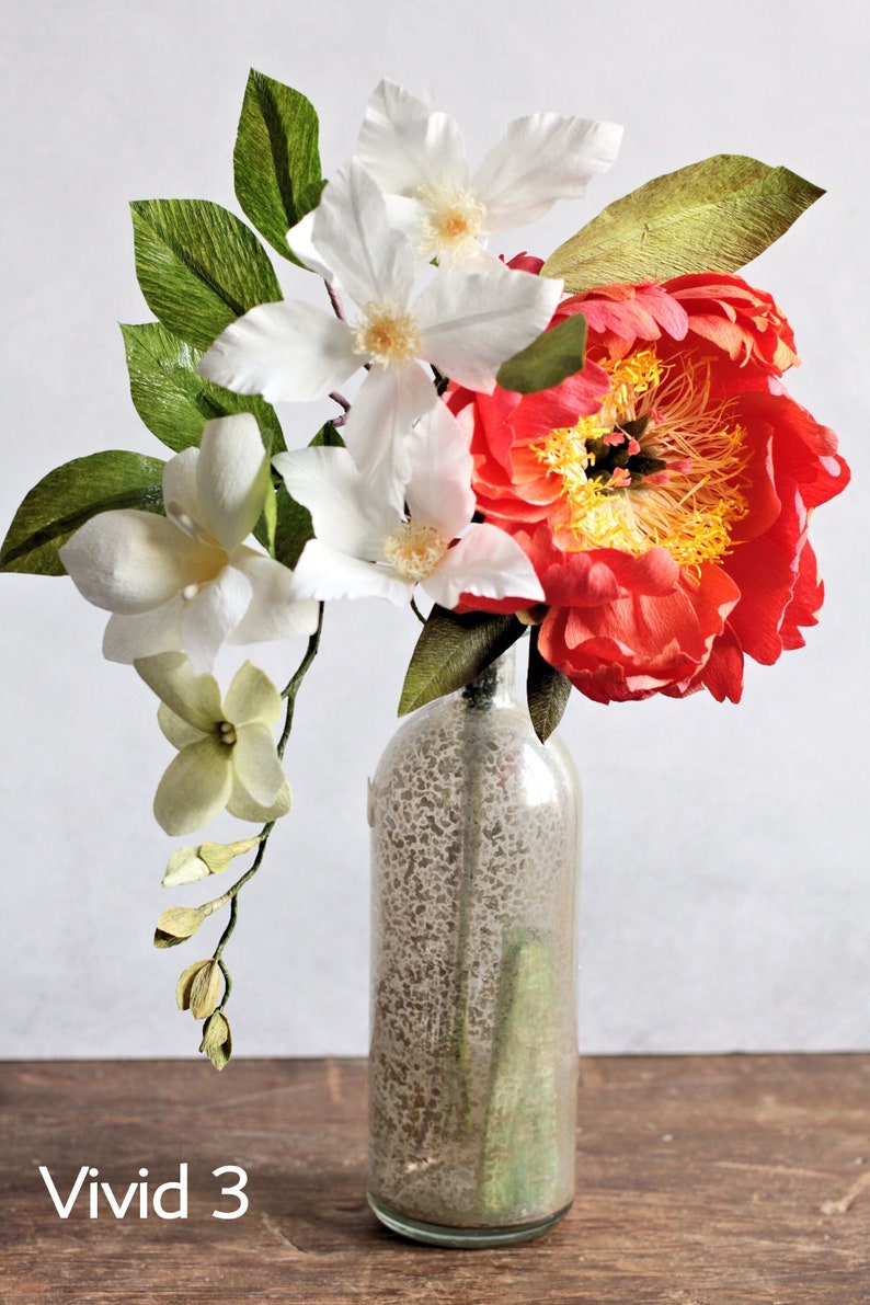 May include: A bouquet of paper flowers in a clear glass bottle with a textured surface. The bouquet includes white clematis, white magnolia, and a large coral-colored peony.