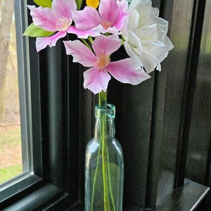 May include: A bouquet of pink and white paper flowers in a clear glass bottle with a copper bottom. The flowers are arranged in a vase on a dark wooden surface. The text "Vivid 15" is visible in the lower left corner of the image.
