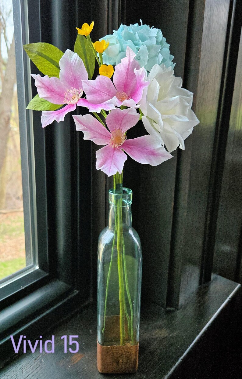 May include: A bouquet of pink and white paper flowers in a clear glass bottle with a copper bottom. The flowers are arranged in a vase on a dark wooden surface. The text "Vivid 15" is visible in the lower left corner of the image.