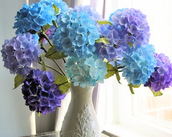 May include: A bouquet of blue and purple hydrangea flowers in a cream-colored vase. The flowers range in shades from light blue to deep purple, with green leaves. The vase has a floral relief pattern.