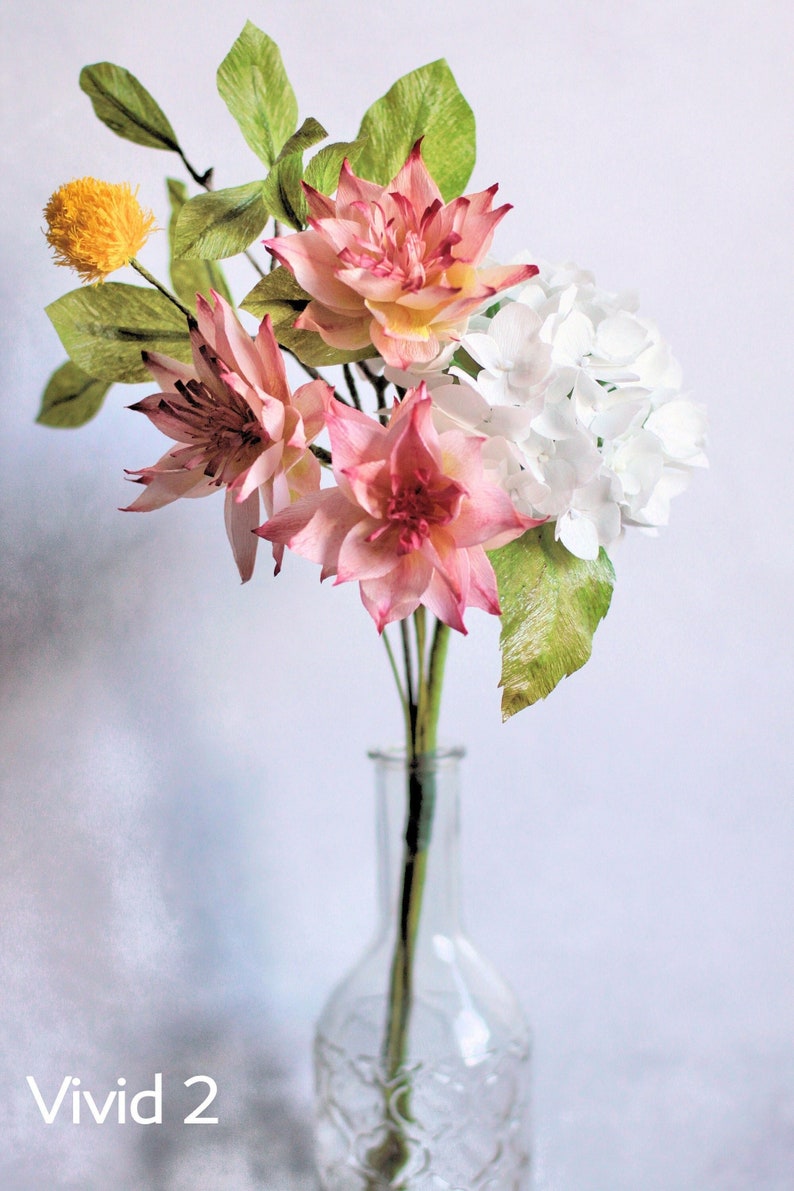 May include: A bouquet of paper flowers in a clear glass vase. The bouquet includes pink and white flowers with green leaves. The flowers are arranged in a vase with a textured surface. The text "Vivid 2" is visible in the bottom left corner of the image.