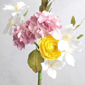 May include: A bouquet of paper flowers in shades of pink, white, and yellow. The bouquet includes a pink hydrangea, white clematis, and a yellow ranunculus. The stems are wrapped in green tape and the text "Vivid 12" is visible at the bottom of the image.