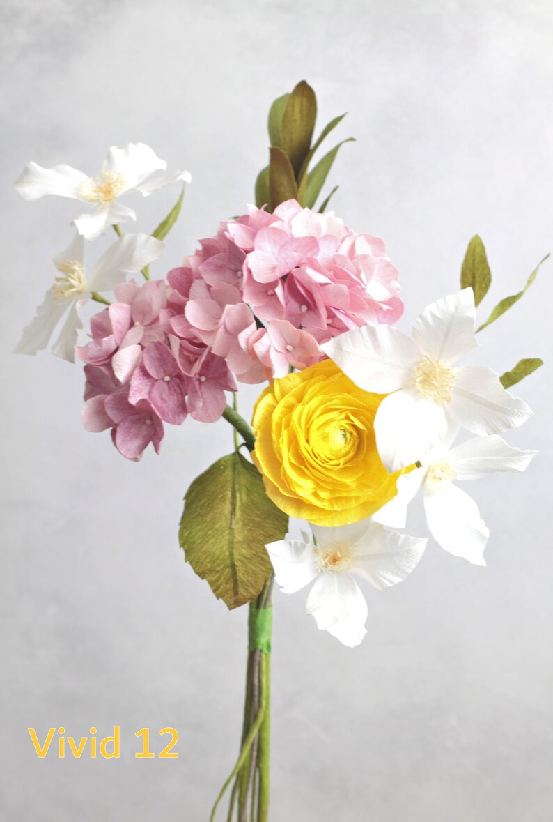 May include: A bouquet of paper flowers in shades of pink, white, and yellow. The bouquet includes a pink hydrangea, white clematis, and a yellow ranunculus. The stems are wrapped in green tape and the text "Vivid 12" is visible at the bottom of the image.
