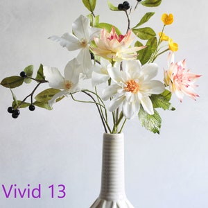 May include: A white ceramic vase with an intricate design holds a bouquet of paper flowers. The flowers are white, pink, and yellow, and include dahlias, clematis, and other varieties. The vase is decorated with a repeating pattern of leaves and flowers.