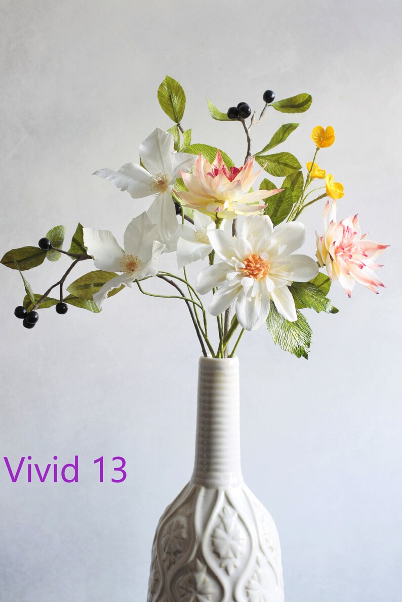 May include: A white ceramic vase with an intricate design holds a bouquet of paper flowers. The flowers are white, pink, and yellow, and include dahlias, clematis, and other varieties. The vase is decorated with a repeating pattern of leaves and flowers.
