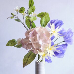 May include: A bouquet of paper flowers in a white vase. The bouquet includes pink hydrangeas, white flowers with yellow centers, and purple flowers. The text "Vivid 14" is visible in the bottom left corner of the image.