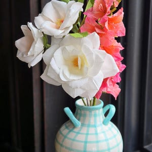 May include: A bouquet of paper flowers in a blue and white checkered vase. The bouquet includes white roses, pink carnations, and pink and white flowers. The vase has a rustic, farmhouse style.