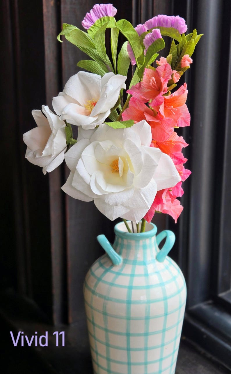 May include: A bouquet of paper flowers in a blue and white checkered vase. The bouquet includes white roses, pink carnations, and pink and white flowers. The vase has a rustic, farmhouse style.