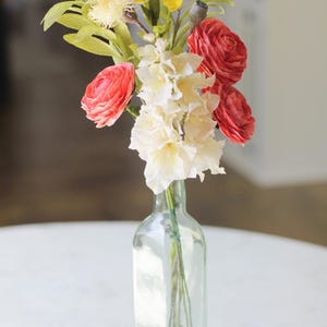 May include: A bouquet of pink and white paper flowers in a clear glass vase with a gold bottom. The vase is sitting on a white table. The text "Vivid 6" is visible in the bottom left corner of the image.
