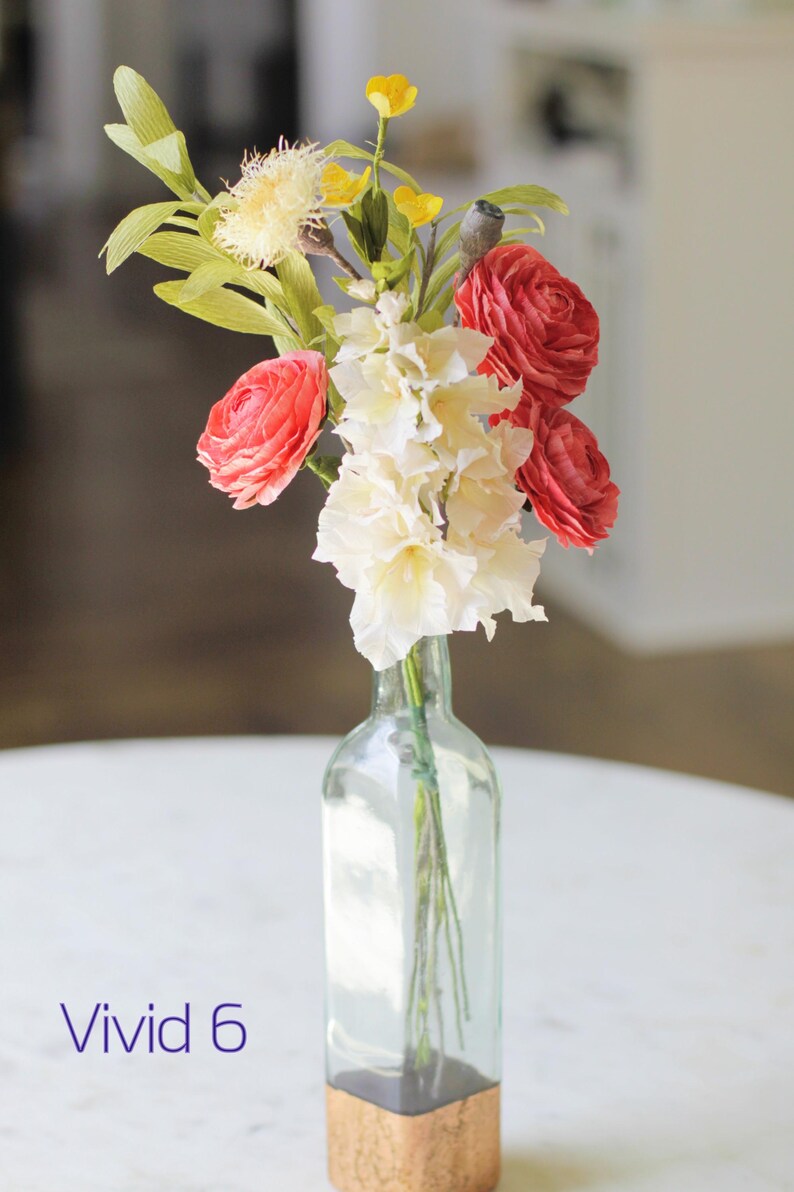 May include: A bouquet of pink and white paper flowers in a clear glass vase with a gold bottom. The vase is sitting on a white table. The text "Vivid 6" is visible in the bottom left corner of the image.