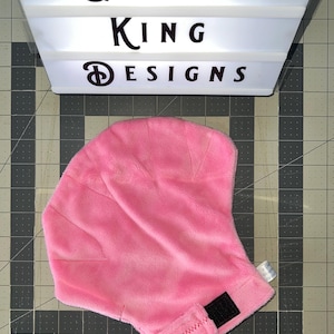 May include: A pink fleece hat with a black velcro closure. The hat is displayed on a gray and black grid surface. Above the hat is a light box with the text "GLIDER KING DESIGNS".