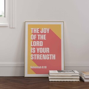 K&ouml;nnte beinhalten: Ein gerahmter Druck mit dem Text "THE JOY OF THE LORD IS YOUR STRENGTH" in Wei&szlig;, vor einem gelben und roten geometrischen Hintergrund. Der Druck wird auf einem Holzboden mit einem Stapel B&uuml;cher in der N&auml;he angezeigt.