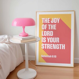 K&ouml;nnte beinhalten: Ein modernes Schlafzimmer mit einem wei&szlig;en Beistelltisch, einer rosa Lampe und zwei B&uuml;chern. Ein gerahmter Druck mit dem Text "THE JOY OF THE LORD IS YOUR STRENGTH" h&auml;ngt an der Wand. Die Lampe hat einen runden Schirm.