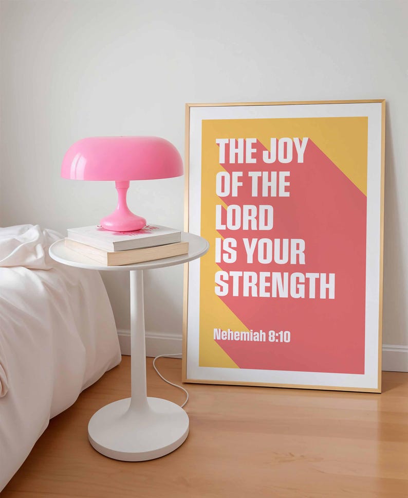 K&ouml;nnte beinhalten: Ein modernes Schlafzimmer mit einem wei&szlig;en Beistelltisch, einer rosa Lampe und zwei B&uuml;chern. Ein gerahmter Druck mit dem Text "THE JOY OF THE LORD IS YOUR STRENGTH" h&auml;ngt an der Wand. Die Lampe hat einen runden Schirm.