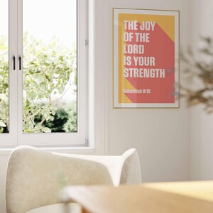 K&ouml;nnte beinhalten: Ein gerahmter Kunstdruck mit dem Text "THE JOY OF THE LORD IS YOUR STRENGTH" in Wei&szlig; auf gelbem und rotem Hintergrund. Der Druck wird an einer wei&szlig;en Wand in der N&auml;he eines Fensters und eines cremefarbenen Stuhls angezeigt.