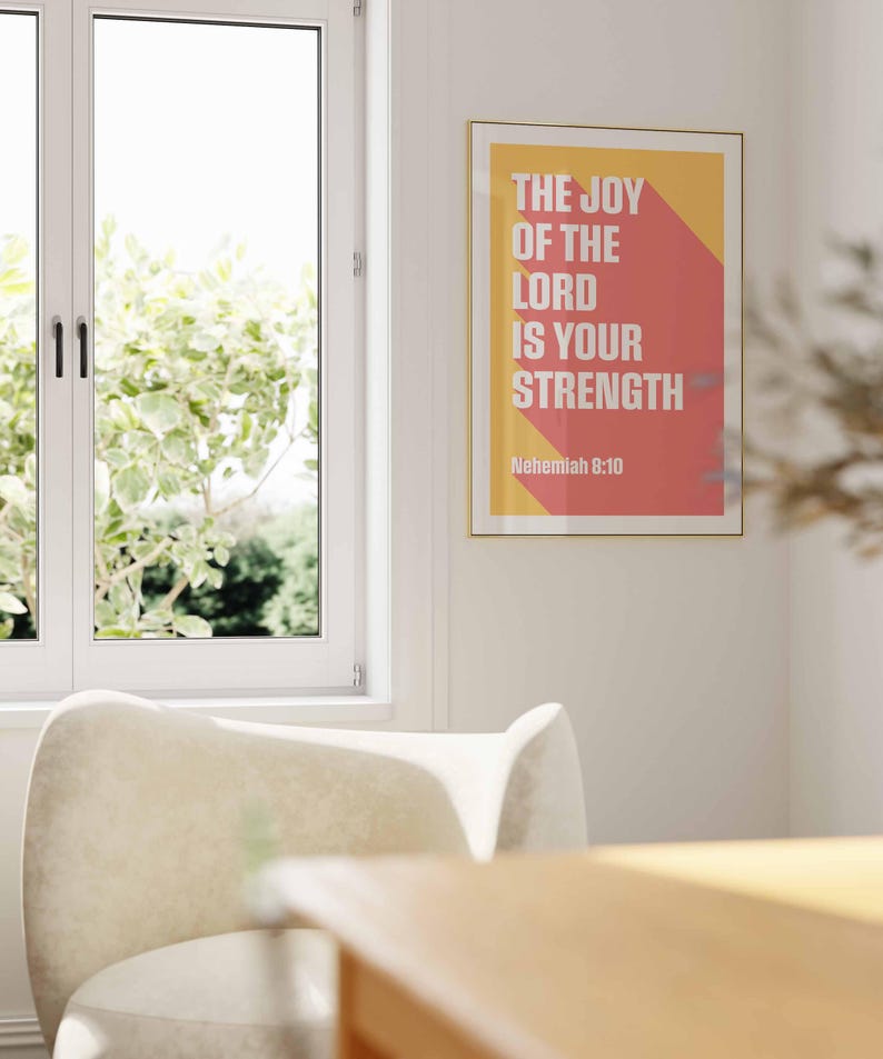 K&ouml;nnte beinhalten: Ein gerahmter Kunstdruck mit dem Text "THE JOY OF THE LORD IS YOUR STRENGTH" in Wei&szlig; auf gelbem und rotem Hintergrund. Der Druck wird an einer wei&szlig;en Wand in der N&auml;he eines Fensters und eines cremefarbenen Stuhls angezeigt.