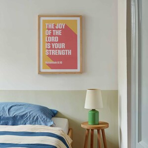 K&ouml;nnte beinhalten: Ein gerahmter Druck mit dem Text "THE JOY OF THE LORD IS YOUR STRENGTH" in wei&szlig;en Buchstaben auf rotem und gelbem Hintergrund. Der Druck h&auml;ngt &uuml;ber einem Bett mit blauem Kissen und einer gr&uuml;nen Lampe auf einem Holzschemel.