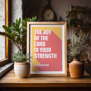 K&ouml;nnte beinhalten: Ein gerahmter Druck mit dem Text "THE JOY OF THE LORD IS YOUR STRENGTH" in Wei&szlig;, vor einem gelb-roten Hintergrund. Der Druck befindet sich in einem Holzrahmen und steht auf einem Holztisch mit Pflanzen.