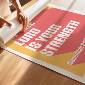 K&ouml;nnte beinhalten: Ein Poster mit dem Text "THE LORD IS YOUR STRENGTH" in Wei&szlig;, vor einem korallenfarbenen und gelben Hintergrund. Der Bibelvers "Nehemia 8:10" wird ebenfalls angezeigt. Das Poster wird auf einer Holzoberfl&auml;che ausgerollt.