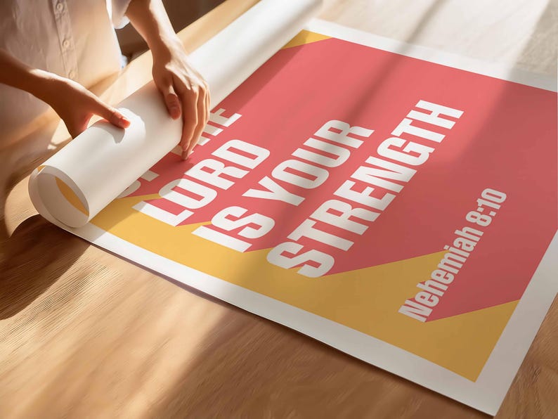 K&ouml;nnte beinhalten: Ein Poster mit dem Text "THE LORD IS YOUR STRENGTH" in Wei&szlig;, vor einem korallenfarbenen und gelben Hintergrund. Der Bibelvers "Nehemia 8:10" wird ebenfalls angezeigt. Das Poster wird auf einer Holzoberfl&auml;che ausgerollt.