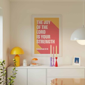 K&ouml;nnte beinhalten: Ein gerahmter Druck mit dem Text "THE JOY OF THE LORD IS YOUR STRENGTH" in Wei&szlig; und einem roten und gelben Hintergrund. Der Druck befindet sich &uuml;ber einem wei&szlig;en Schrank mit einer gelben Lampe, Kerzen und anderen dekorativen Gegenst&auml;nden. Eine wei&szlig;e Pendelleuchte h&auml;ngt von der Decke.