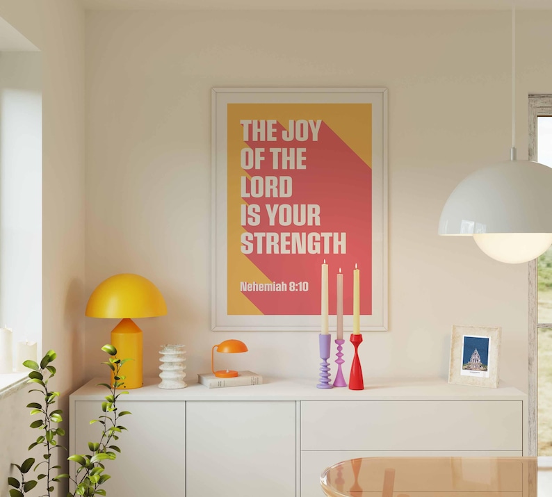 K&ouml;nnte beinhalten: Ein gerahmter Druck mit dem Text "THE JOY OF THE LORD IS YOUR STRENGTH" in Wei&szlig; und einem roten und gelben Hintergrund. Der Druck befindet sich &uuml;ber einem wei&szlig;en Schrank mit einer gelben Lampe, Kerzen und anderen dekorativen Gegenst&auml;nden. Eine wei&szlig;e Pendelleuchte h&auml;ngt von der Decke.