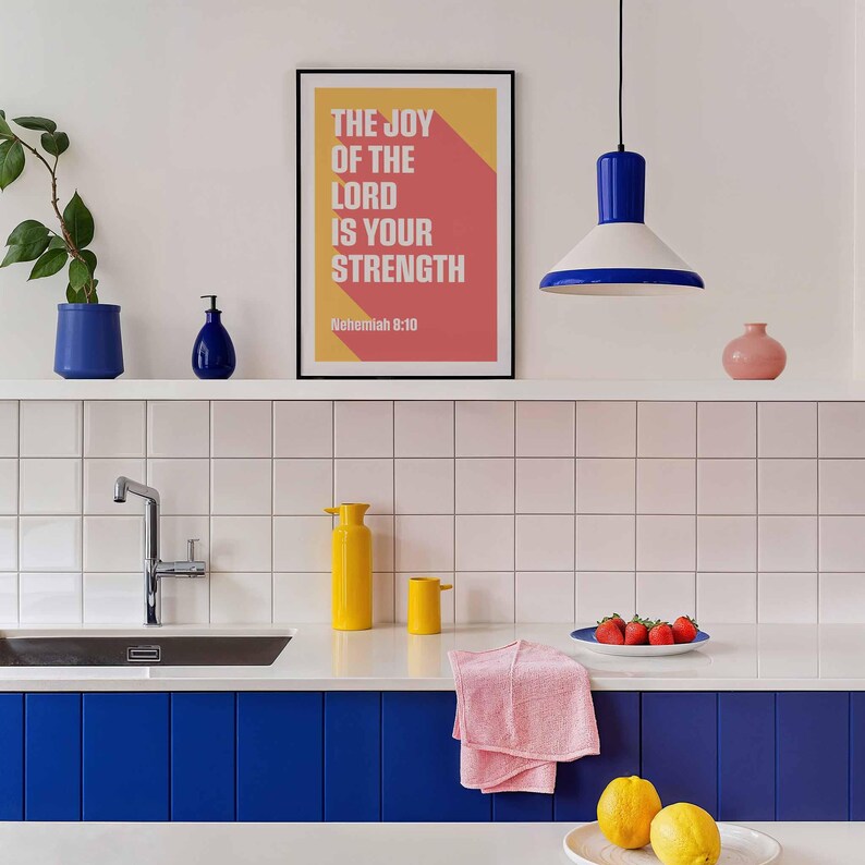K&ouml;nnte beinhalten: Moderne K&uuml;chenszene mit wei&szlig;er Arbeitsplatte, blauen Schr&auml;nken und gefliestem Fliesenspiegel. Ein gerahmter Druck mit dem Text "THE JOY OF THE LORD IS YOUR STRENGTH" h&auml;ngt &uuml;ber der Theke. Eine gelbe Vase und ein rosa Handtuch liegen auf der Theke.