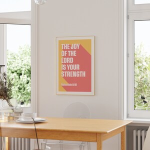 K&ouml;nnte beinhalten: Ein gerahmter Druck mit dem Text "THE JOY OF THE LORD IS YOUR STRENGTH" in Wei&szlig;, vor einem roten und gelben Hintergrund. Der Druck wird in einem Esszimmer an einer wei&szlig;en Wand mit einem Holztisch und St&uuml;hlen pr&auml;sentiert.