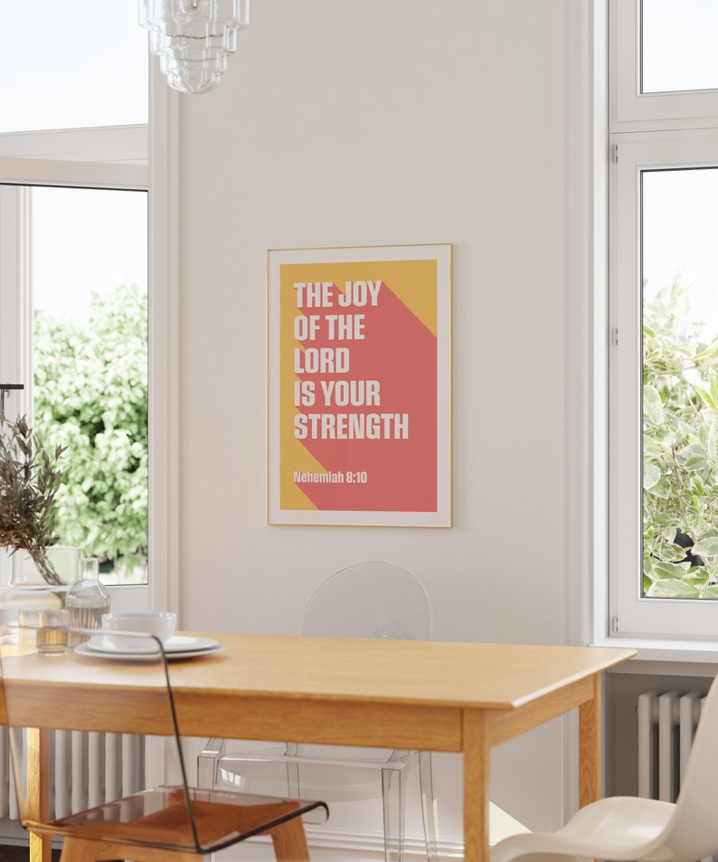 K&ouml;nnte beinhalten: Ein gerahmter Druck mit dem Text "THE JOY OF THE LORD IS YOUR STRENGTH" in Wei&szlig;, vor einem roten und gelben Hintergrund. Der Druck wird in einem Esszimmer an einer wei&szlig;en Wand mit einem Holztisch und St&uuml;hlen pr&auml;sentiert.