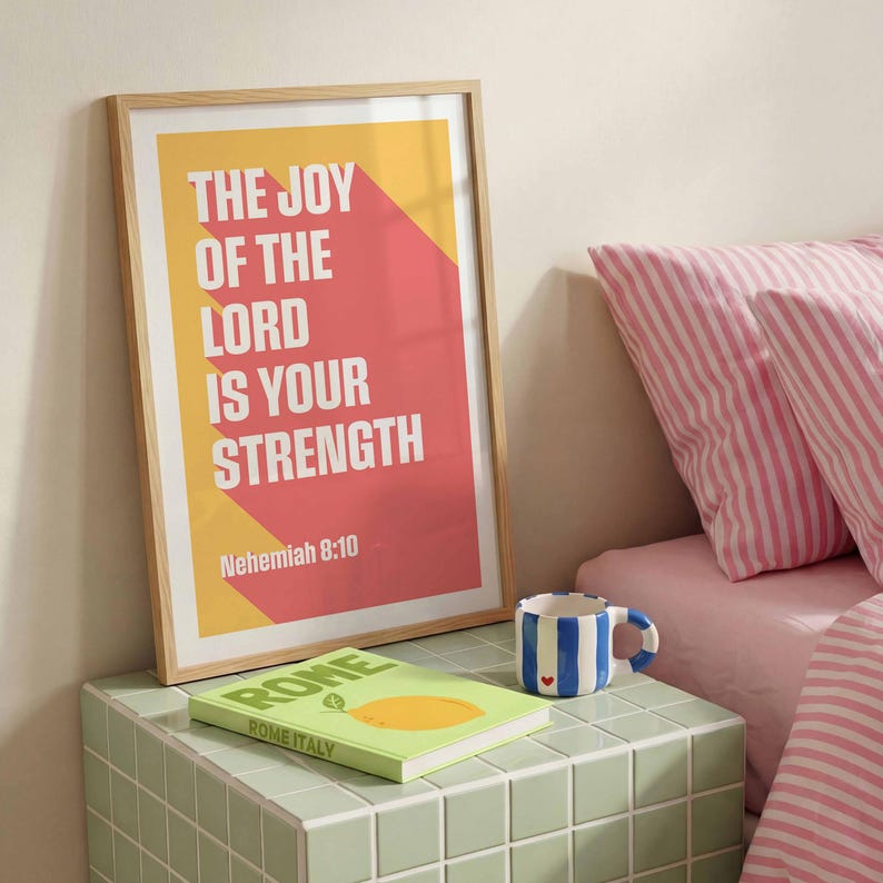 K&ouml;nnte beinhalten: Ein gerahmtes Poster mit dem Text "THE JOY OF THE LORD IS YOUR STRENGTH" in Wei&szlig;, vor gelben und roten geometrischen Formen. Ein Buch mit dem Titel "ROME ITALY" und eine blau-wei&szlig; gestreifte Tasse mit rotem Herz liegen auf einer gr&uuml;n gefliesten Oberfl&auml;che.