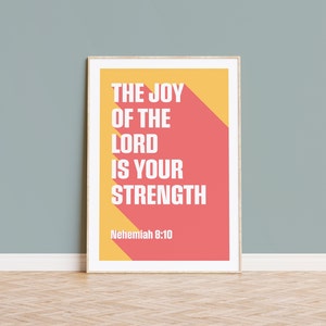 K&ouml;nnte beinhalten: Gerahmter Kunstdruck mit dem wei&szlig;en Text "THE JOY OF THE LORD IS YOUR STRENGTH" auf einem korallenroten und gelben Hintergrund. Das Zitat stammt aus Nehemia 8:10. Der Druck wird an einer hellblauen Wand gezeigt.