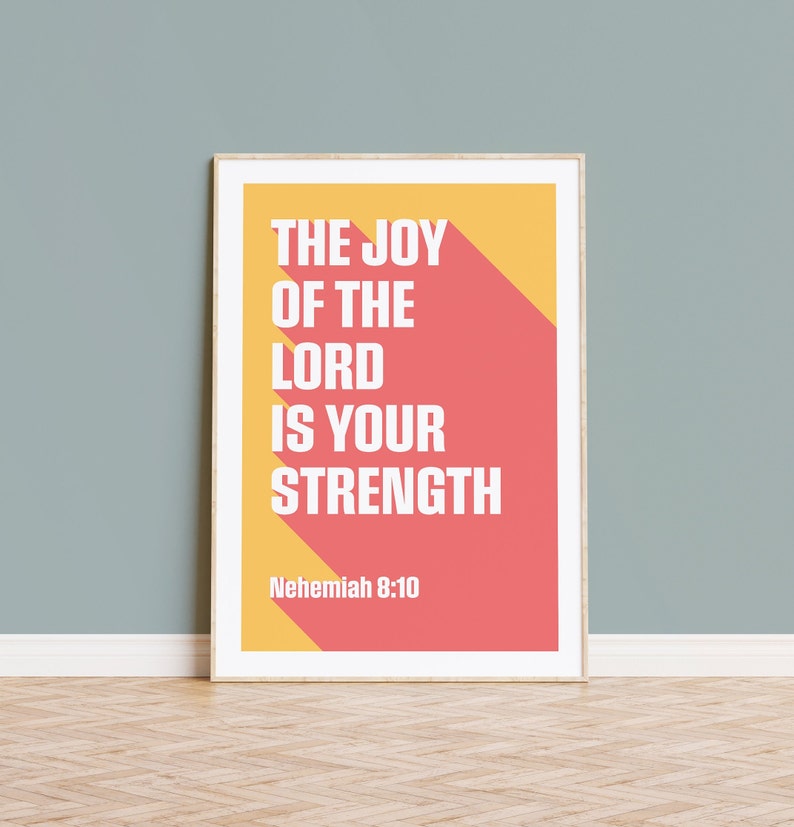 K&ouml;nnte beinhalten: Gerahmter Kunstdruck mit dem wei&szlig;en Text "THE JOY OF THE LORD IS YOUR STRENGTH" auf einem korallenroten und gelben Hintergrund. Das Zitat stammt aus Nehemia 8:10. Der Druck wird an einer hellblauen Wand gezeigt.