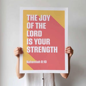 K&ouml;nnte beinhalten: Ein Poster mit dem Text "THE JOY OF THE LORD IS YOUR STRENGTH" in Wei&szlig;, vor einem roten und gelben Hintergrund. Der Text "Nehemiah 8:10" befindet sich unten. Das Poster wird von einer Person gehalten.